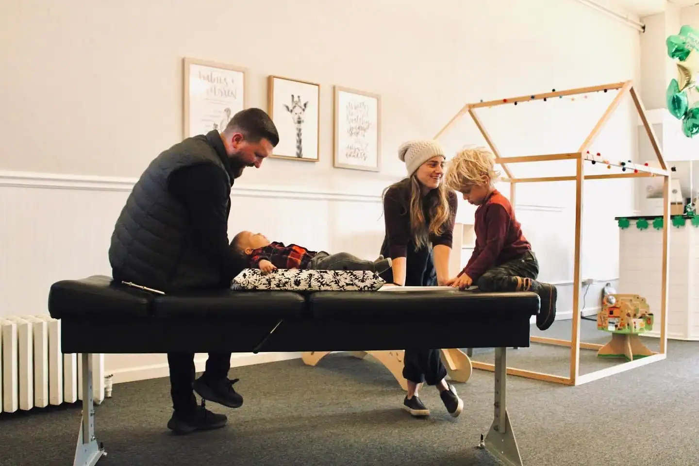 Photo of a chiropractor sitting on a bench with a baby lying down next to a mom and her young son