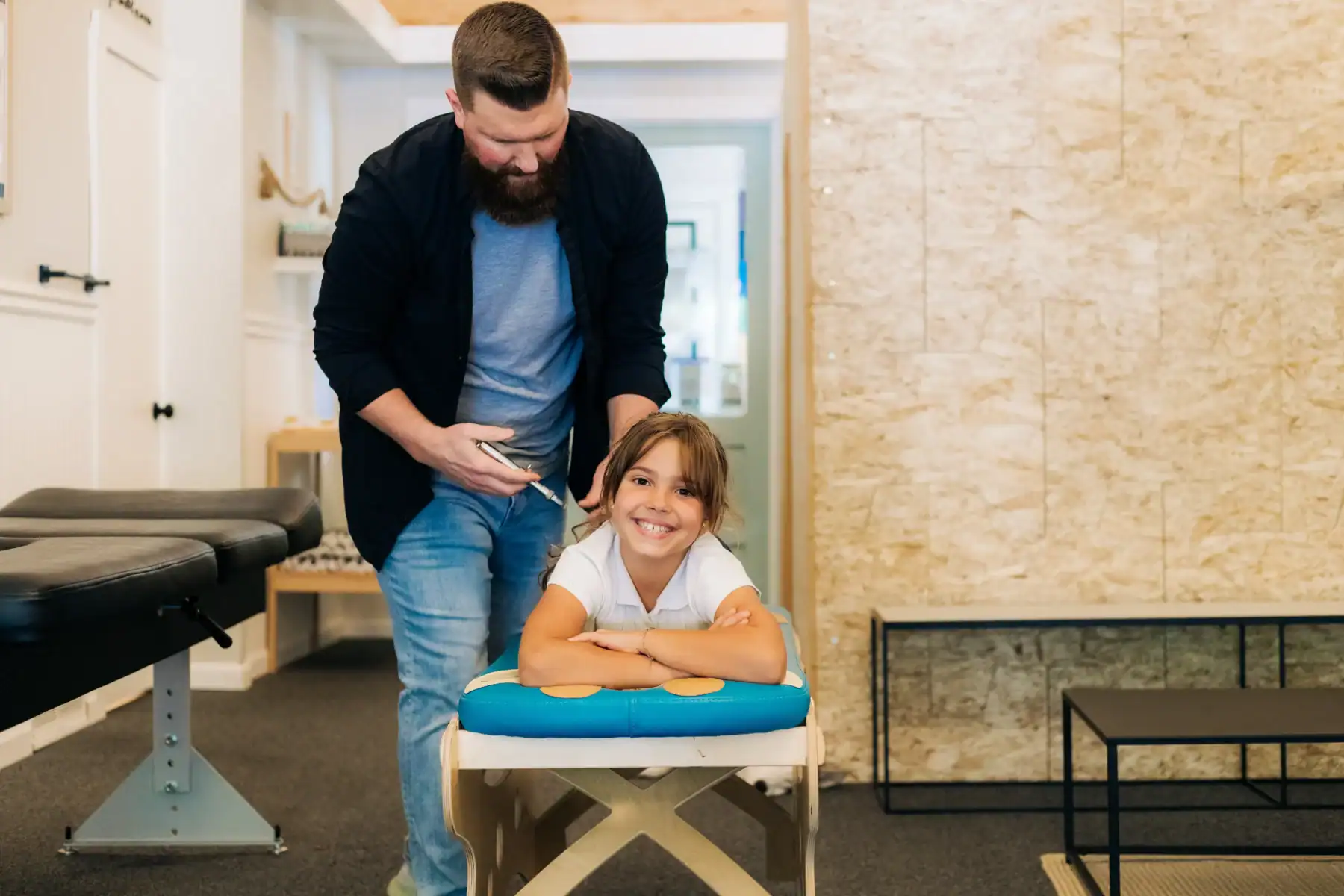 Photo of a chiropractor using a tool on a young girl's back while she lies on a bench smiling