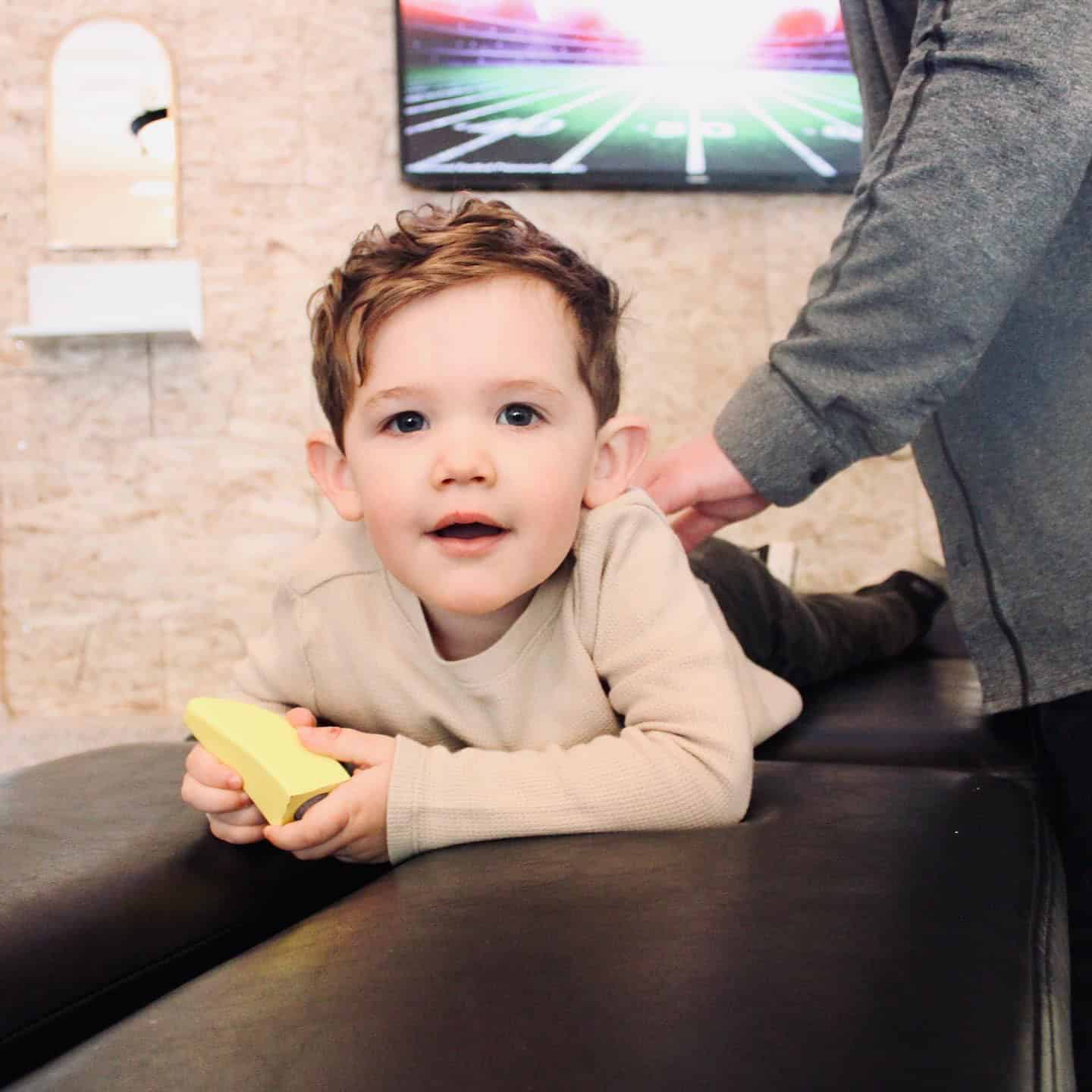 Photo of a young boy lying on his stomach on a bench while a chiropractor adjusts his back