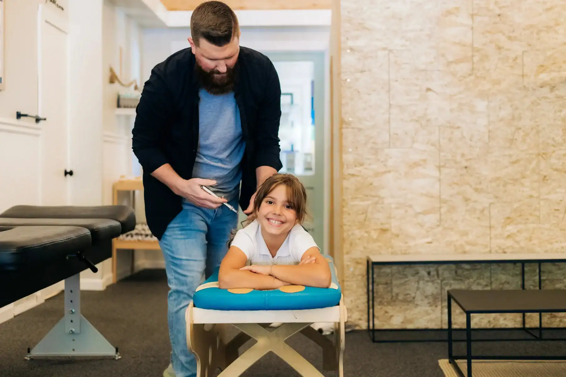photo of what is a pediatric chiropractor using a tool on a young girl s back while she lies on a bench smiling