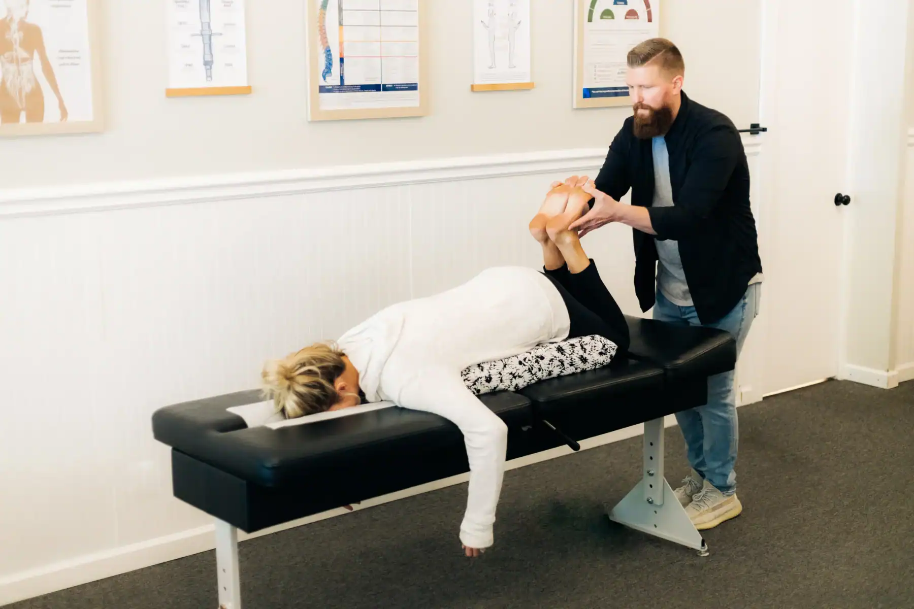 photo of a chiropractor lifting the legs of a female patient as she lies on her stomach on a black bench