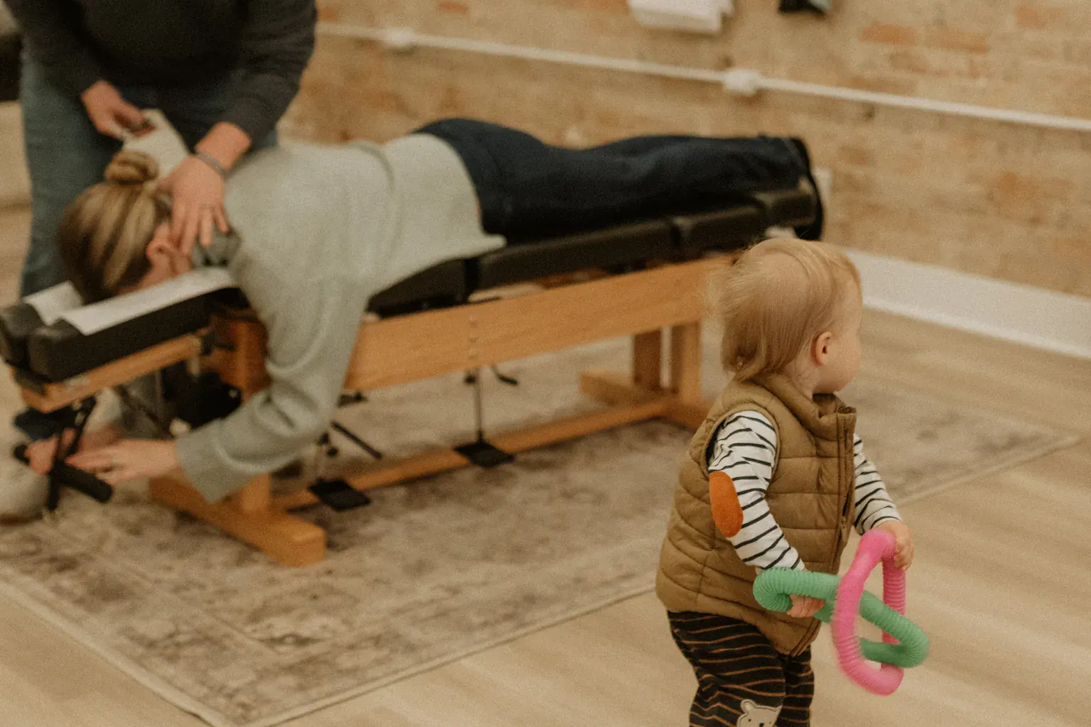 photo of a toddler holding a toy while a chiropractor adjusts a patient in the background